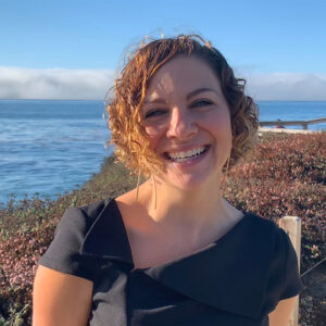 A young white woman with curly hair smiles broadly at the camera. She wears a dark shirt and stands on a cliffside, with coastal scrub, clouds, and the Pacific Ocean in the background.
