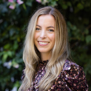 Photograph of a young blonde woman smiling at the camera. She wears a dark floral shirt and stands against a background of dark green foliage.