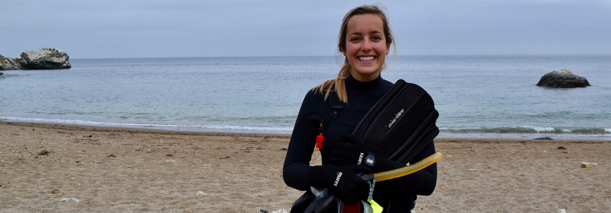 Photograph of a young white woman wearing a wetsuit. She stands on a beach with gray clouds in the background.