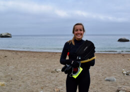 Photograph of a young white woman wearing a wetsuit. She stands on a beach with gray clouds in the background.