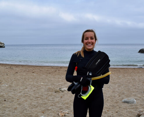 Photograph of a young white woman wearing a wetsuit. She stands on a beach with gray clouds in the background.