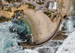 Aerial view of the La Jolla Children's Pool, with white-capped waves crashing against the dark gray stone of a seawall. The dark brown shapes of sea lions dot the protected beach.