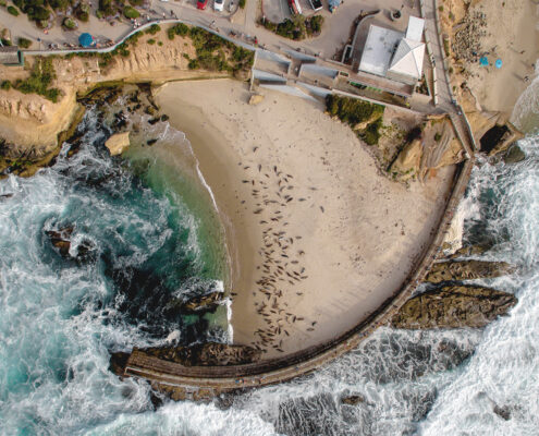 Aerial view of the La Jolla Children's Pool, with white-capped waves crashing against the dark gray stone of a seawall. The dark brown shapes of sea lions dot the protected beach.