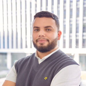 Photograph of a young bearded Latine man smiling at the camera. He wears a white polo shirt and gray sweater with a yellow rectangular pin.