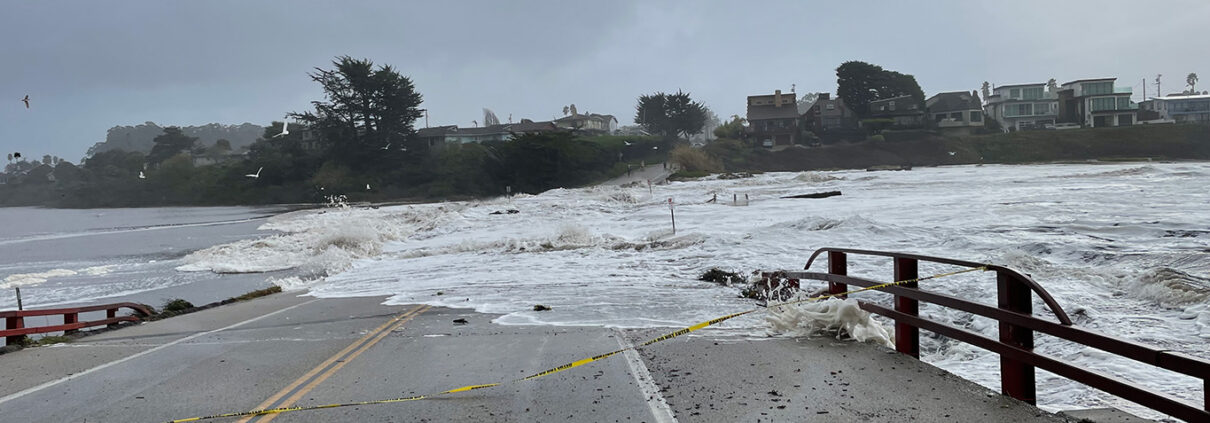 The viewer looks down a bridge, most of which is hidden underwater. Waves wash across the road and trees loom against a gray sky on the opposite side of the flood.