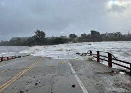 The viewer looks down a bridge, most of which is hidden underwater. Waves wash across the road and trees loom against a gray sky on the opposite side of the flood.