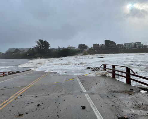 The viewer looks down a bridge, most of which is hidden underwater. Waves wash across the road and trees loom against a gray sky on the opposite side of the flood.