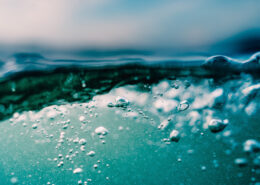 Bubbles rise erratically towards the rippling surface of water. Particulates are visible below the surface, and faint clouds smudge the sky above.