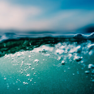 Bubbles rise erratically towards the rippling surface of water. Particulates are visible below the surface, and faint clouds smudge the sky above.