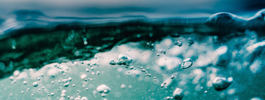 Bubbles rise erratically towards the rippling surface of water. Particulates are visible below the surface, and faint clouds smudge the sky above.