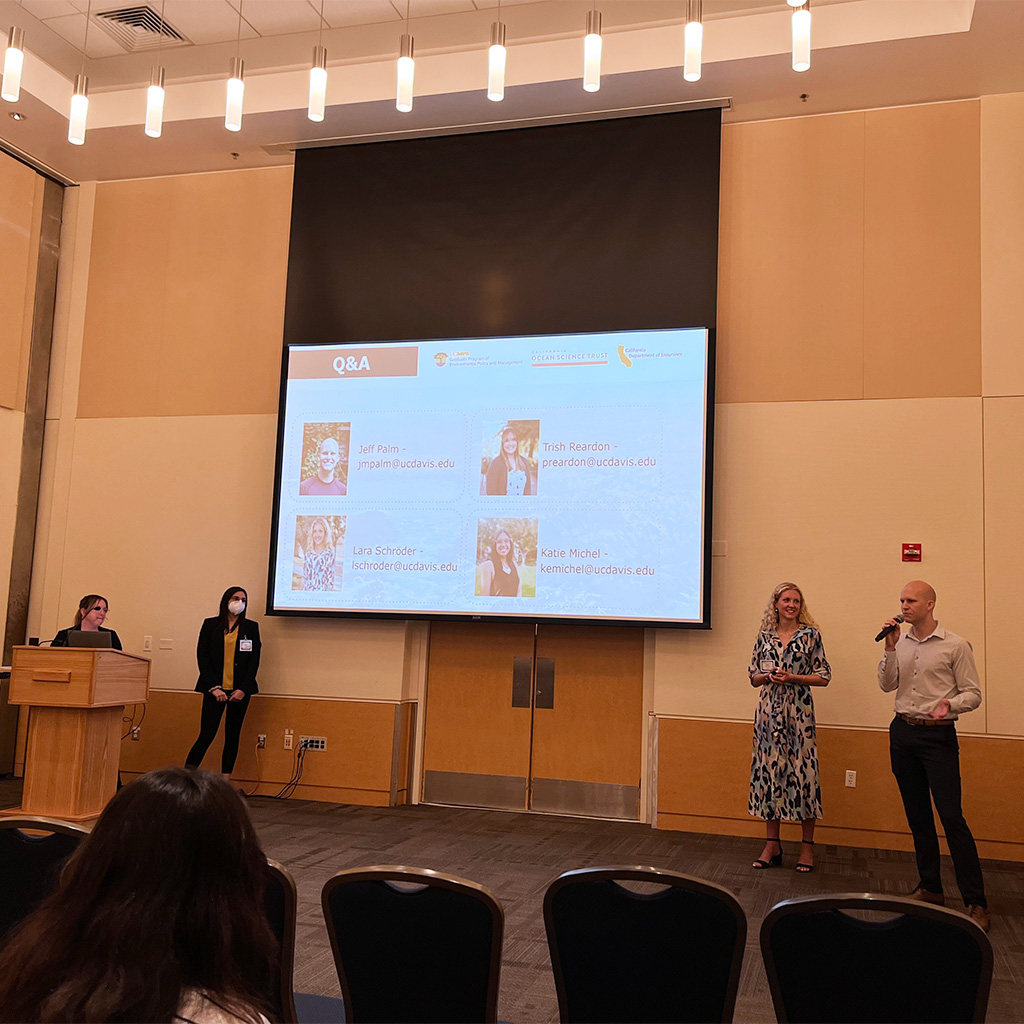 Four graduate students stand below a screen displaying their names and headshots. The bald white man on the far right is speaking into a microphone while the others watch him.