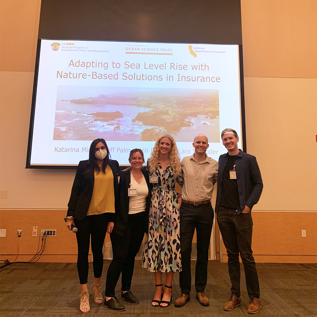 Five people stand in front of their presentation screen and smile at the camera.