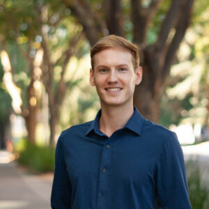 A young white man with blond hair smiles at the camera. He stands in front of a blurry tree and wears a dark blue button-down shirt.