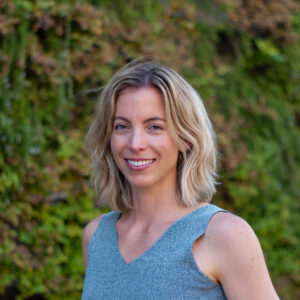 A white woman with shoulder-length blonde hair smiles at the camera. She stands against a soft green background and wears a gray sleeveless shirt.