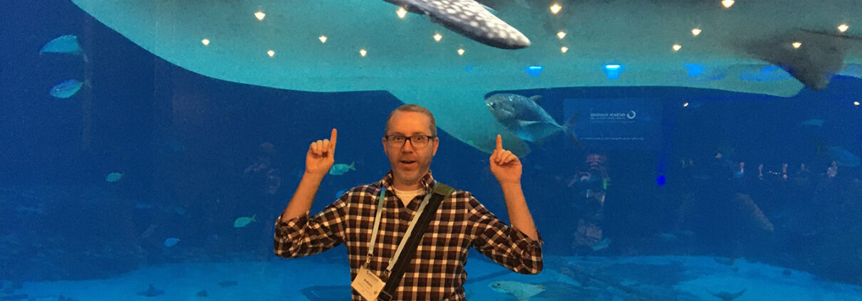 A white man in a plaid shirt points up with both hands. Behind him is a deep blue tank, with a whale shark and pilot fish swimming past just beyond the glass.