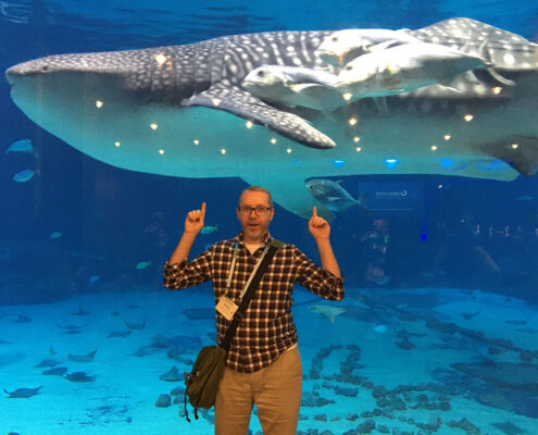 A white man in a plaid shirt points up with both hands. Behind him is a deep blue tank, with a whale shark and pilot fish swimming past just beyond the glass.