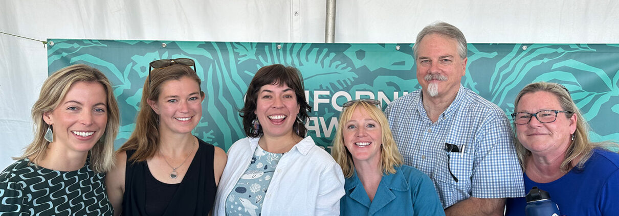 Six people smile at the camera, standing in front of a mostly-hidden banner of seaweed and a white pavilion wall.