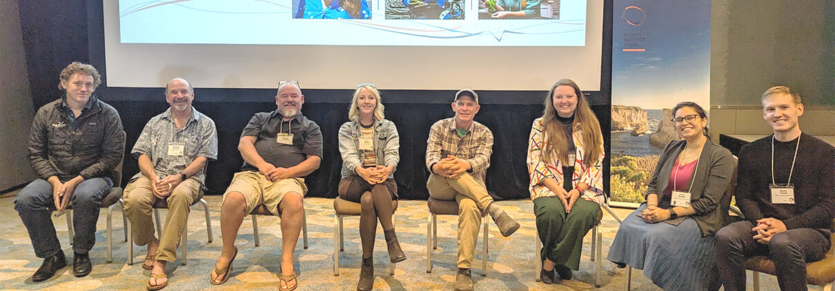 A photograph of eight panelists smiling at the camera. The lower edge of a projector screen is visible behind them.