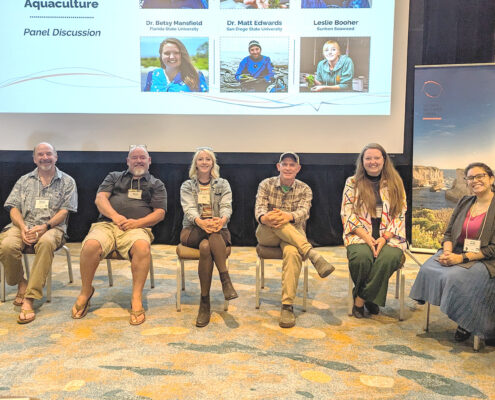 A photograph of eight panelists smiling at the camera. The lower edge of a projector screen is visible behind them.