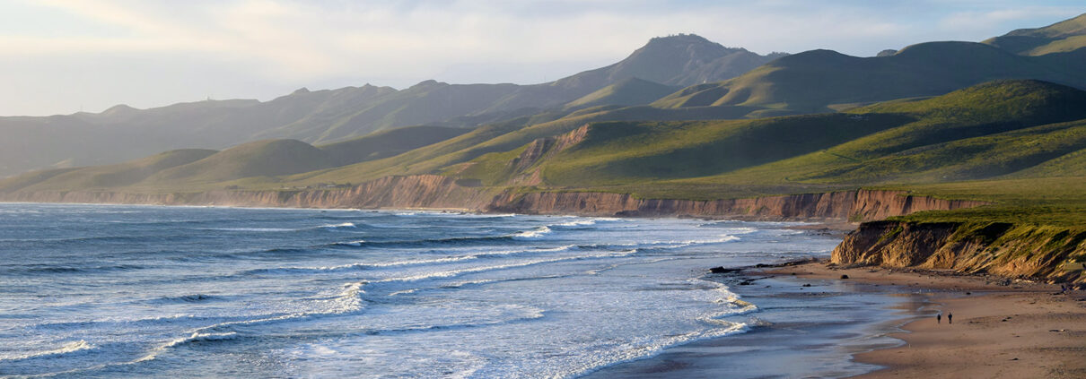 Photograph of the Central California coastline, looking north towards green hills. The wide beach and low whitecaps are tinted gold by the afternoon sun.