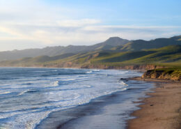 Photograph of the Central California coastline, looking north towards green hills. The wide beach and low whitecaps are tinted gold by the afternoon sun.