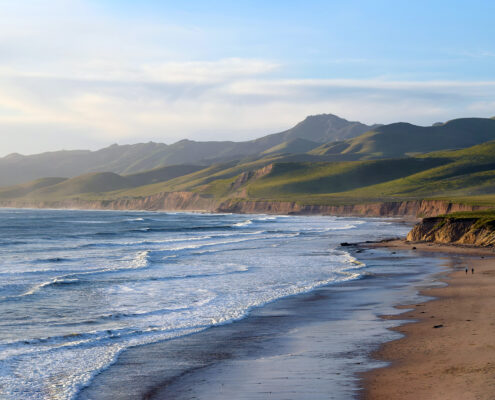 Photograph of the Central California coastline, looking north towards green hills. The wide beach and low whitecaps are tinted gold by the afternoon sun.