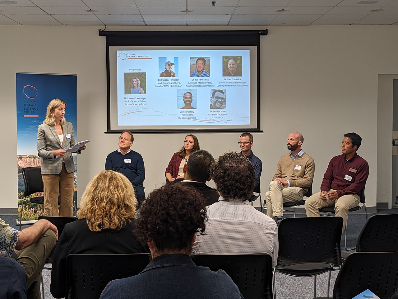 Six people sit and stand at the front of a beige room. A Powerpoint presentation is visible on the screen behind them.