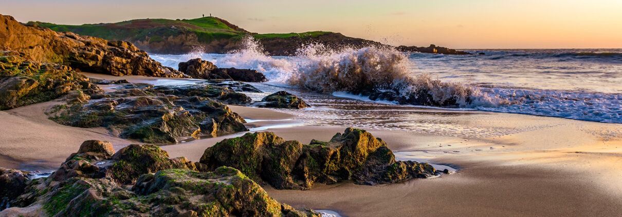 Photograph of Bean Hollow State Beach at sunset; waves crash against rocks, and a small cliff is visible in the background.