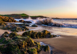 Photograph of Bean Hollow State Beach at sunset; waves crash against rocks, and a small cliff is visible in the background.
