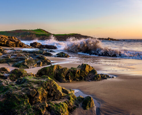 Photograph of Bean Hollow State Beach at sunset; waves crash against rocks, and a small cliff is visible in the background.