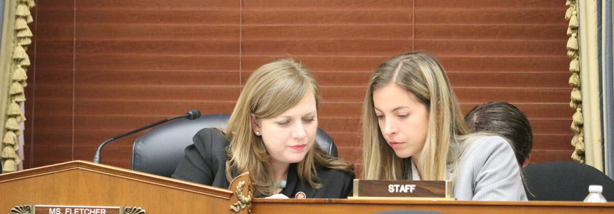 Two blonde women sit behind a desk, looking at something on the desk.