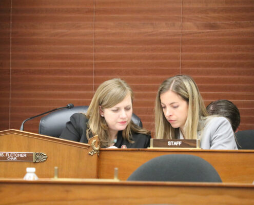 Two blonde women sit behind a desk, looking at something on the desk.