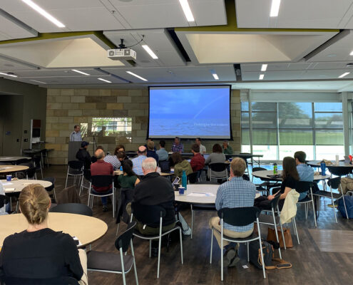 Photograph of a presentation during the May 2024 Fisheries Disaster symposium. Peoples backs are visible as they face the presenter. A large screen can be seen at the far end of the room.