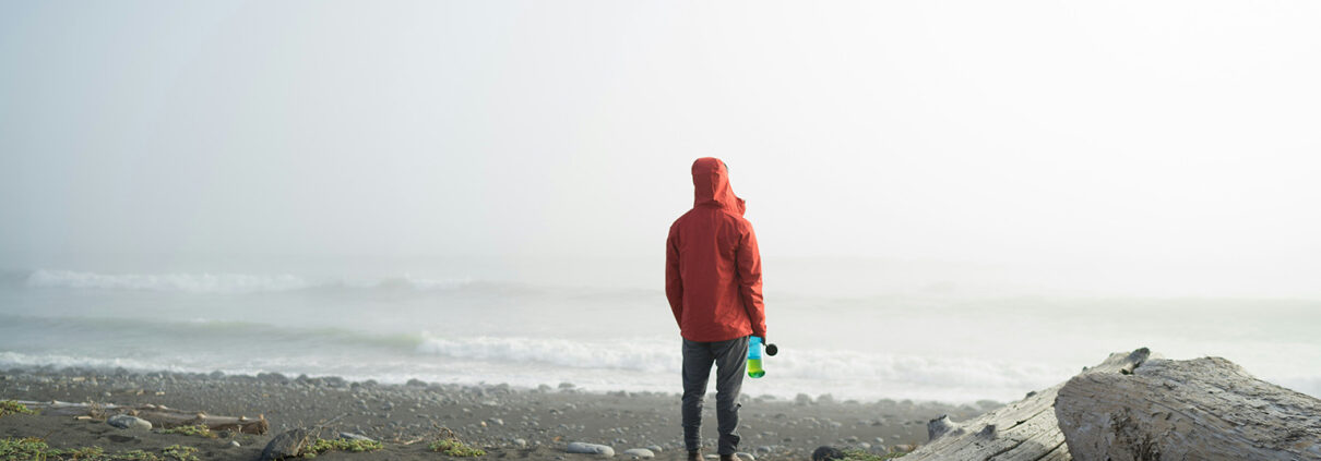 Photograph of a person in a red jacket standing with their back to the camera. A driftwood log is visible next to them, and the sea recedes into fog in behind them.