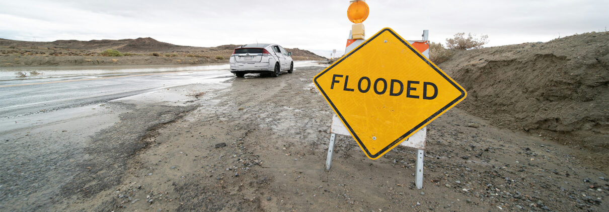 A car parked near a flooded roadway. A large yellow sign in the foreground says "FLOODED"