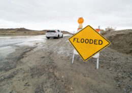 A car parked near a flooded roadway. A large yellow sign in the foreground says "FLOODED"