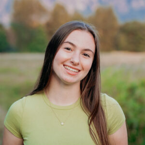 A young white woman with brown hair smiles at the camera. She wears a light green T-shirt and stands in front of a field, with trees visible in the background.