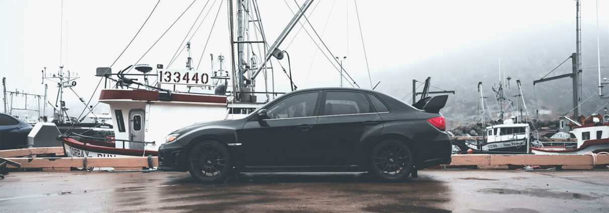 A black car parked on a foggy dock. Fishing boats are visible in the harbor behind the car.
