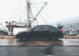 A black car parked on a foggy dock. Fishing boats are visible in the harbor behind the car.